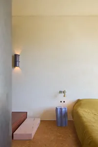 A minimalist bedroom featuring a concrete wall, a yellow bedspread, and a pink wooden bench.