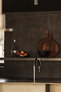 Minimalist kitchen with a concrete backsplash, featuring a stainless steel faucet, a bowl of oranges, and a carafe.