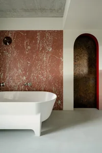 A minimalist bathroom features a red marble wall, a white bathtub, and concrete-like flooring.