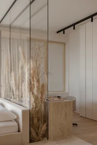 A modern minimalist bedroom featuring a wooden bed frame, glass closet doors, and fabric curtains with pampas grass plant.
