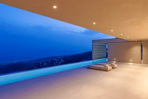 Modern pool deck with concrete walls and a rooftop infinity pool overlooking a night-time landscape with illuminated houses a