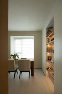 A minimalist dining room featuring a wooden table and beige chairs against a white wall with a large window providing natural