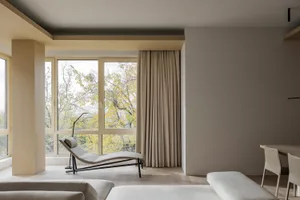 A minimalist living room with neutral fabrics and wood elements, featuring a lounge chair, sofa, and large windows with a vie