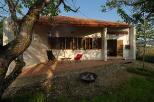 A cozy cottage with a brick patio, two chairs, and a fire pit in front of a white-washed concrete cottage with a red-tiled ro