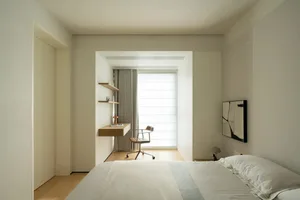 A minimalist bedroom with a white bed, a small wooden desk, and a window letting in natural light.