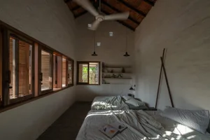 Bedroom with a minimalist design, featuring textured plaster walls, wooden furniture, and natural light from large windows.