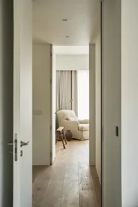 A minimalist residential room with a wooden floor, a beige armchair, and a small wooden stool.