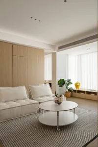 Minimalist modern living room with wood furniture and white accents, featuring open shelving and a large window.