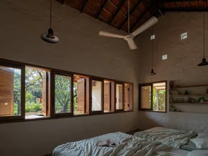 A minimalist bedroom featuring wooden window shutters and a bed with an open book, set against a rustic, high-ceilinged wall.