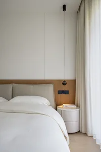 Modern minimalist bedroom featuring a white bed with grey headboard, wooden flooring, and sheer white curtains.