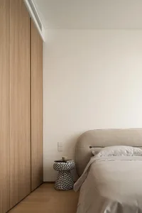 Minimalist bedroom with light wood flooring, a beige upholstered bed, and a patterned side table with a mug and book.