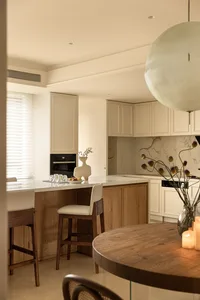 A minimalist residential kitchen with marble countertops and wooden furniture, featuring a round dining table and bar stools.
