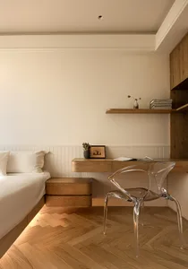 A modern bedroom features a wooden desk with a clear plastic chair, a neatly made bed, and a shelf with decorative items.