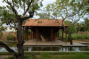 A wooden traditional house garden shelter with a thatched roof, surrounded by greenery and a pond.