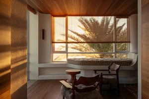 A cozy window seat with a cushioned bench and a wooden table in a white room with wooden walls and floor.