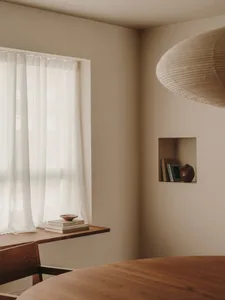 A minimalist interior with a wooden shelf and white sheer curtains, featuring a suspended lamp and a niche with books and a v