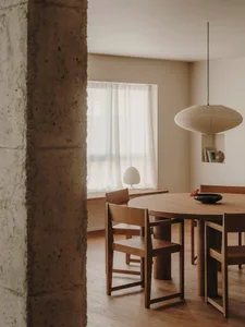 A minimalist kitchen features a wooden table and chairs, a large window with sheer curtains, and a modern pendant lamp.
