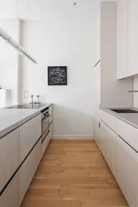 A minimalist white kitchen with a wooden floor, stainless steel appliances, and concrete countertops.