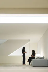 Two people conversing in a minimalist living space with concrete walls and a grey sofa.