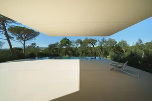 A modern residential exterior featuring a concrete floor and ceiling, with an infinity pool and surrounding trees.