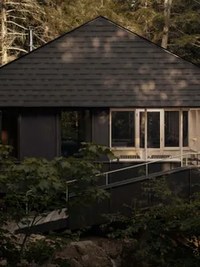 A black-shingled cabin situated along a forested edge, featuring large windows and a metal chimney.