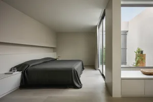 A minimalist bedroom featuring concrete flooring and walls, with a large bed covered in dark sheets and a nightstand with a g