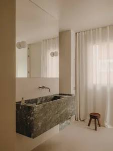 Minimalist bathroom interior with a large, dark marble sink, wall-mounted faucet, and floor-to-ceiling white curtains.