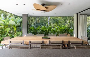 Modern dining room with a wooden table and chairs surrounded by greenery and large windows.