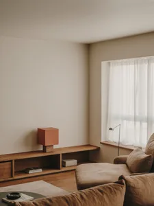 A modern minimalist living room featuring a wooden sofa and table with a red square lamp on a wooden shelf, next to a window 