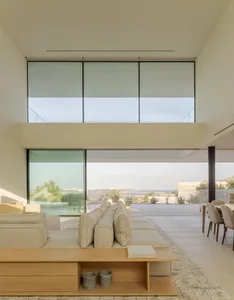 Modern, minimalist interior with beige upholstered furniture, large windows with glass and beige blinds, and a view of an out