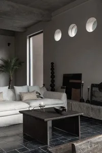 A modern living room with a minimalist design, featuring a concrete floor and walls, large windows, and a cozy seating area.