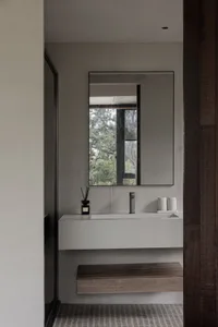 Minimalist bathroom featuring a sleek concrete vanity with a wooden bench, a large mirror, and a window overlooking greenery.