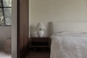 A modern minimalist bedroom with a white fabric bedspread and a minimalist bedside table with a white shade lamp.