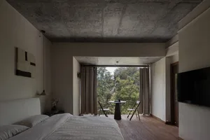 A minimalist bedroom with concrete ceiling, wooden flooring, and views of greenery outside through large sliding glass doors.