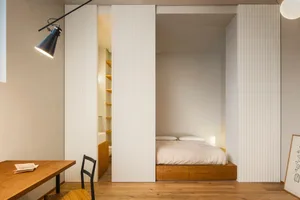 A minimalist bedroom featuring a wooden platform bed with white bedding, surrounded by white textured panels and minimalist l