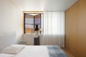 A person standing in front of a window with wooden shutters and blinds in a modern minimalist bedroom setting.