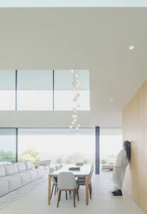 A minimalist living room with large windows and concrete walls, featuring a white sectional sofa, wooden dining table, and mo