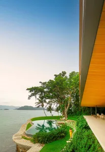 A modern waterfront residence featuring a wooden deck, concrete patio, and lush greenery.