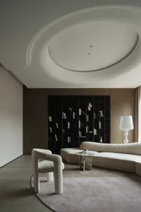 A modern living room with neutral-toned decor, featuring a white sculptural chair, a curved sofa, and a bookshelf filled with
