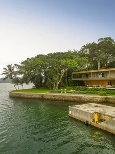 Modern waterfront house with a pool, green lawn, and surrounding trees.
