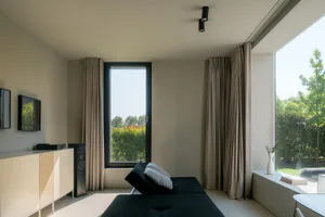 A minimalist living room featuring a black sofa, white drapes, and large windows.