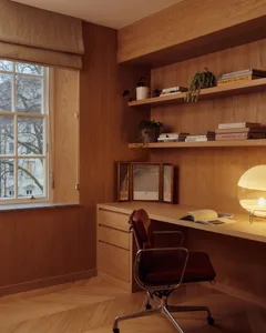 A minimalist office space featuring a wood desk and chair, with shelves containing books and plants, and a window providing n
