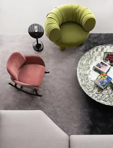 A modern living room featuring a green upholstered armchair, a pink rocking chair, and a round glass table with books and fru