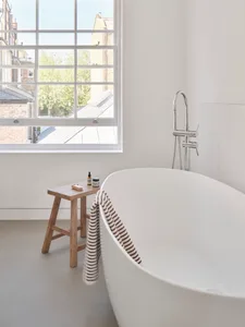 A modern white bathroom with a freestanding ceramic tub and a stool with bottles and a sponge on top.