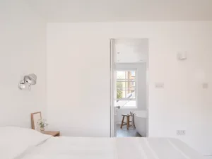 A minimalist bedroom with white walls, bed, and bathroom in the background, featuring a white bathtub and wooden stool.