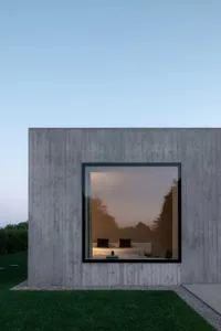 Modern, minimalist concrete box house with a large window reflecting trees and the interior bedroom.