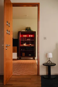 A modern living room with a wooden interior, featuring a door, a cabinet with red lighting and glassware, and a side table wi