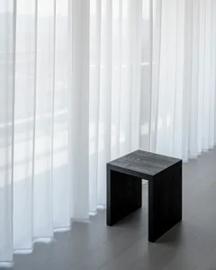 A minimalist studio featuring a black wooden stool and sheer white curtains.