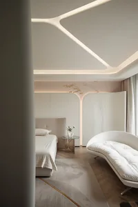 Minimalist bedroom with a beige bed and a curved white sofa, featuring a glass bedside table with a vase of flowers.