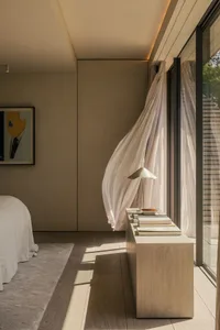 Minimalist bedroom interior with a bed, a window with sheer curtains, and a nightstand with a lamp and books.